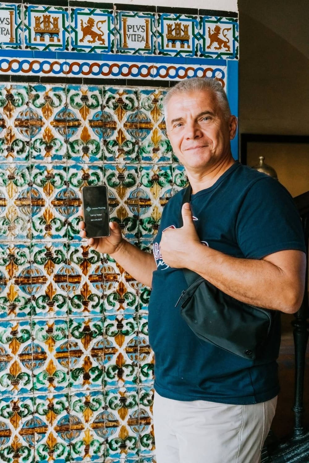 The owner of Leonov Flooring giving a thumbs-up and holding a phone with the company's number in front of a historic Spanish tile wall