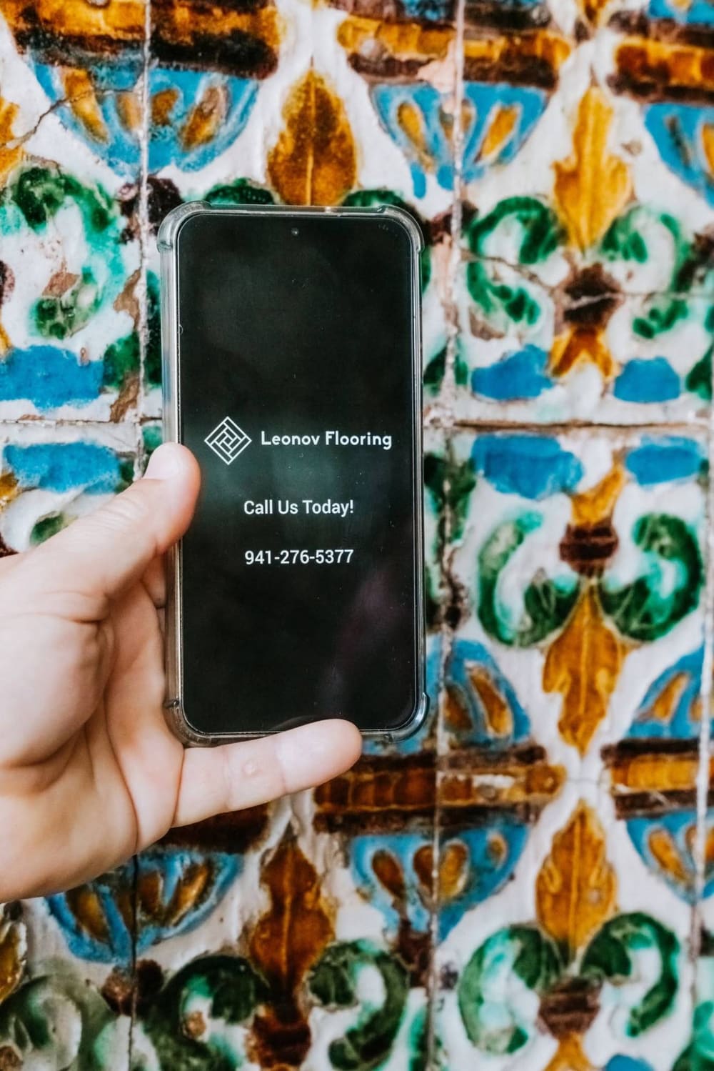 A phone showing the Leonov Flooring logo and phone number held up against colorful hand-painted Spanish tiles